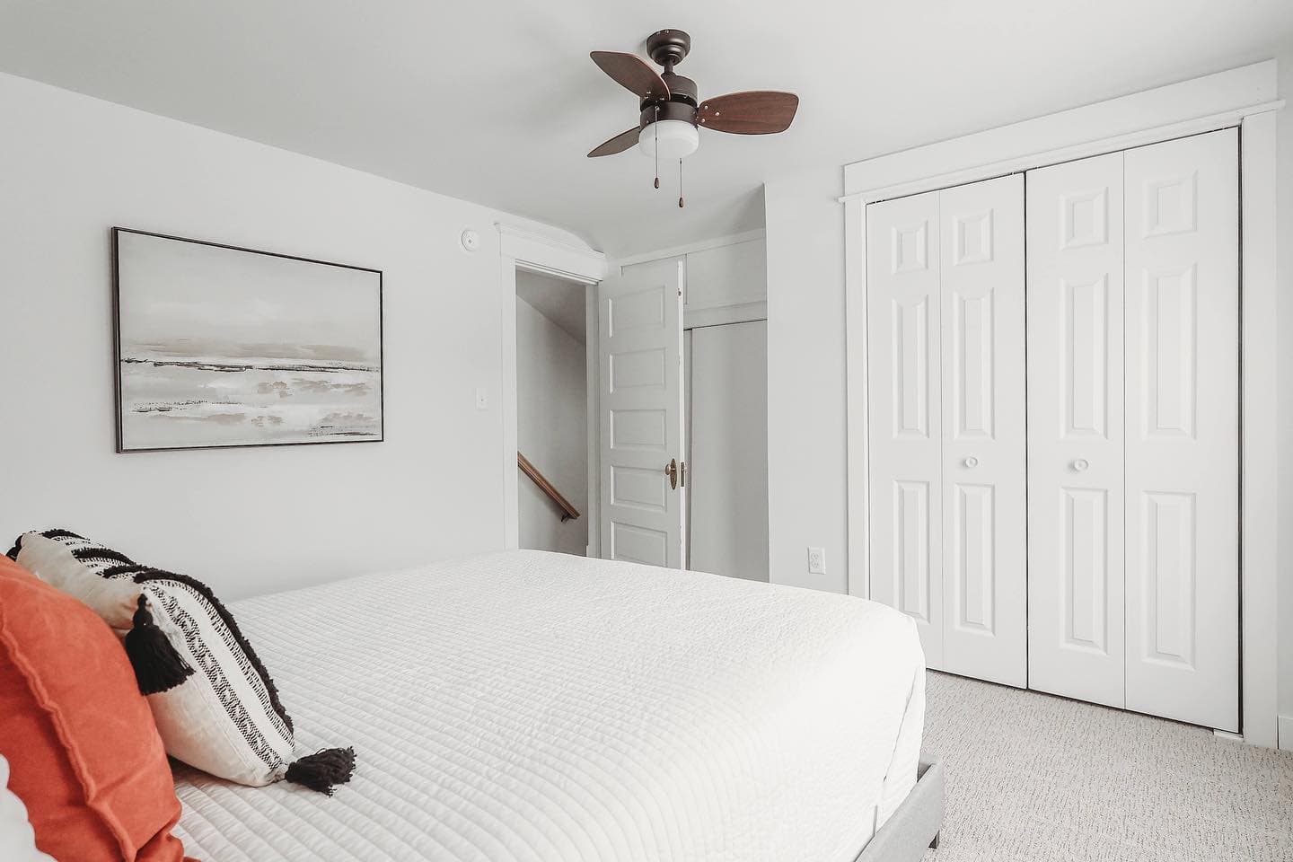 Modern white bedroom with textured bedding, orange accent pillow, abstract art, and dark ceiling fan.