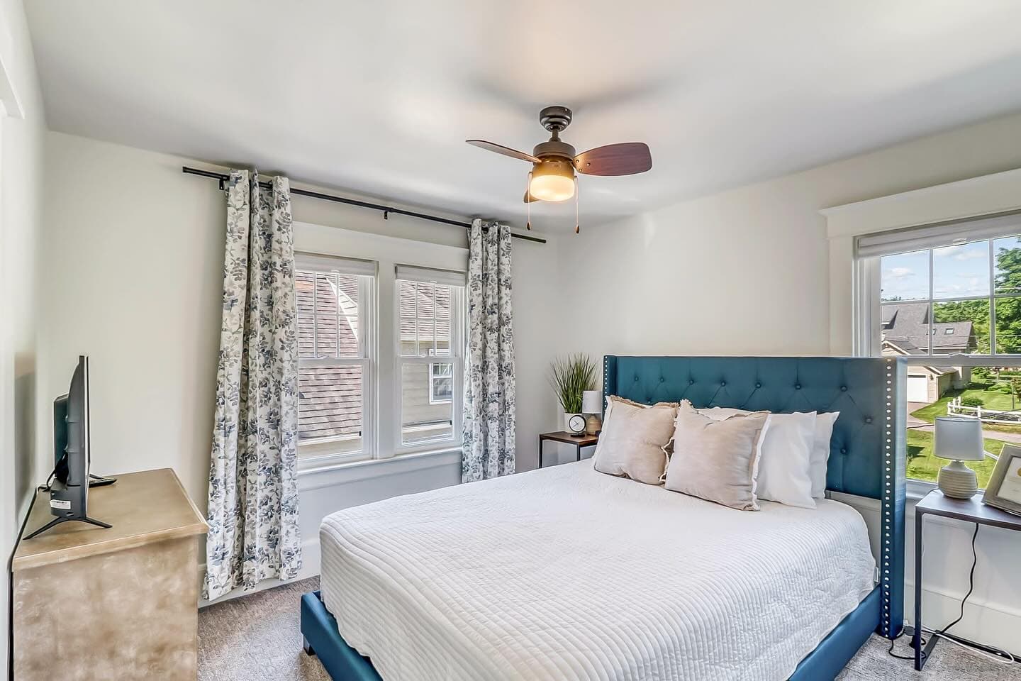 Bright bedroom with a blue tufted headboard, white bedding, and floral patterned window curtains.