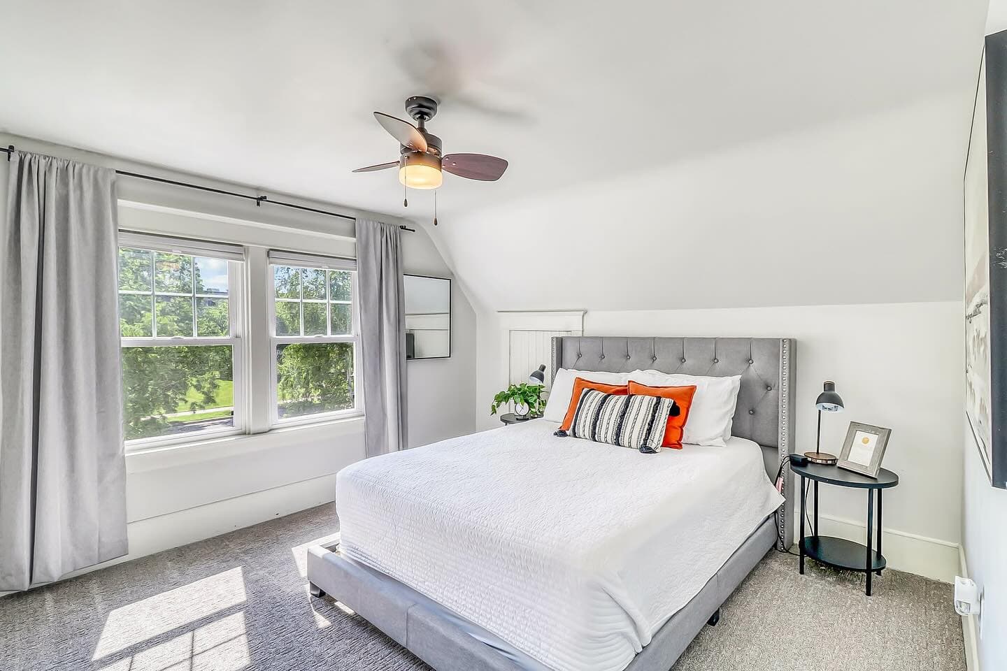 Bright bedroom with a grey tufted bed, white bedding, orange pillows, and large sunlit windows.