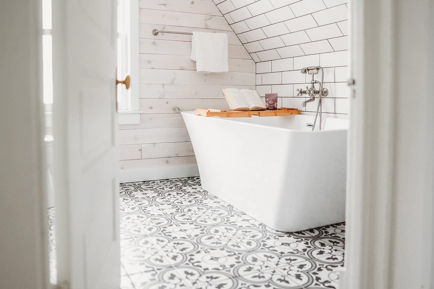 White freestanding bathtub with a wooden tray and book on ornate patterned floor tiles.