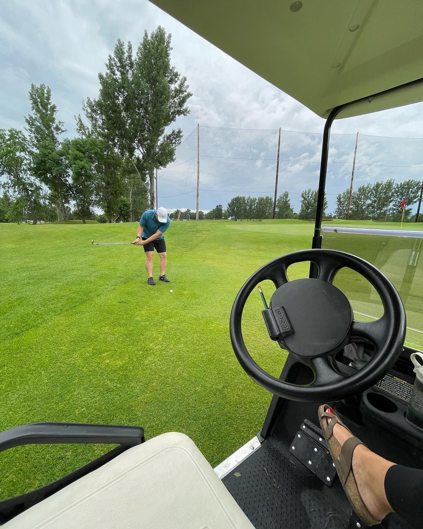 Man swinging a golf club on a green fairway, viewed from inside a golf cart.