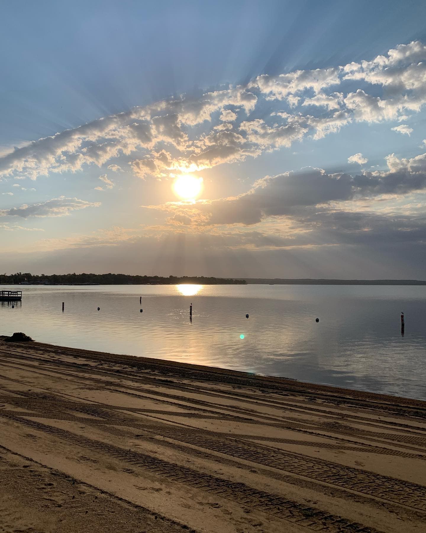 Bright sunbeams pierce through clouds above a calm lake and a sandy, tire-tracked beach.