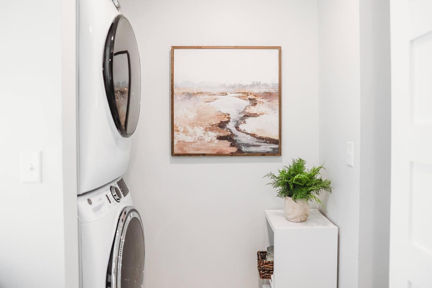 Stacked white washer and dryer next to framed landscape art and a potted plant.