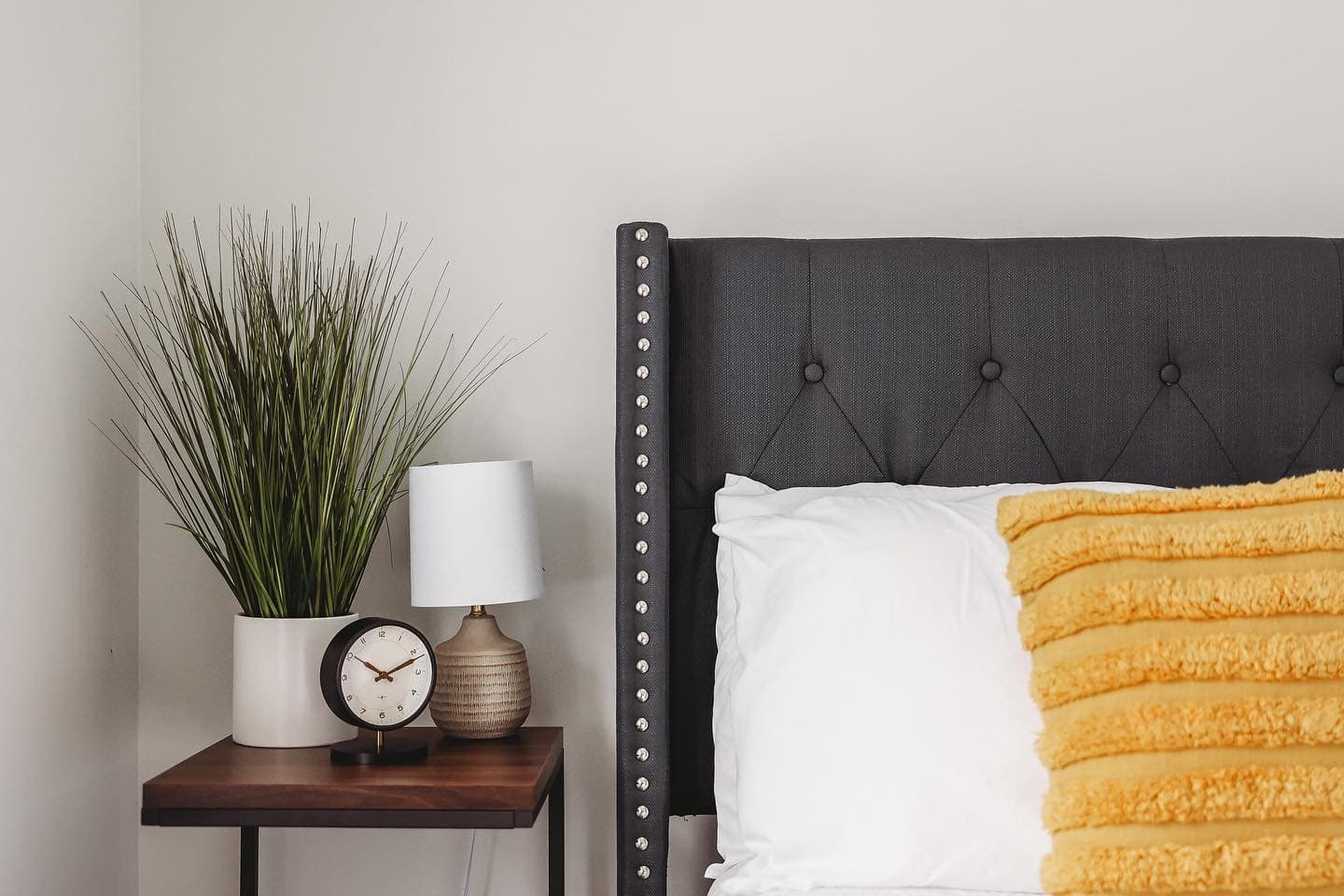 Dark gray tufted headboard with yellow pillow next to a nightstand with clock and plant.