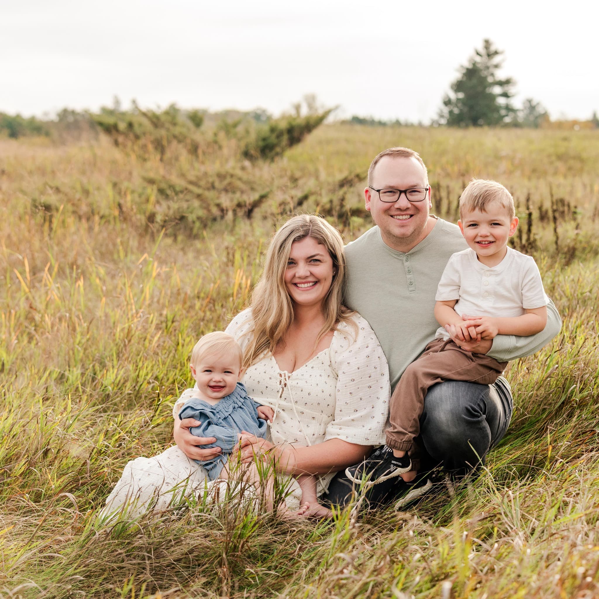Smiling parents with their toddler son and baby daughter posing in a tall grassy field.