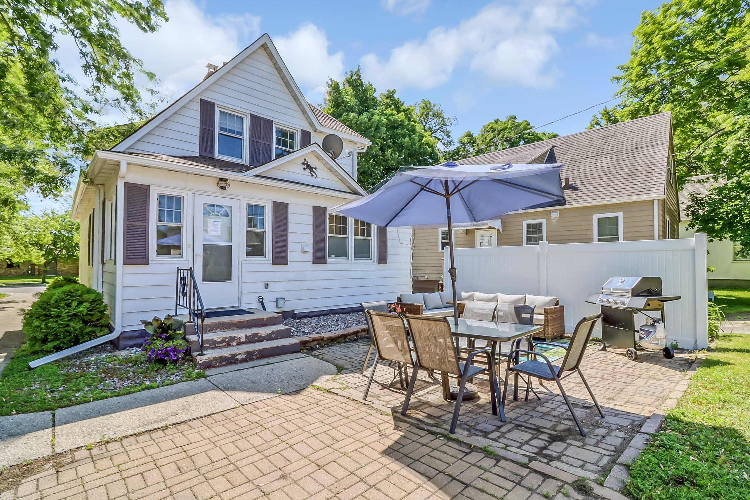 White house with brick patio, dining set under blue umbrella, lounge seating, and grill.