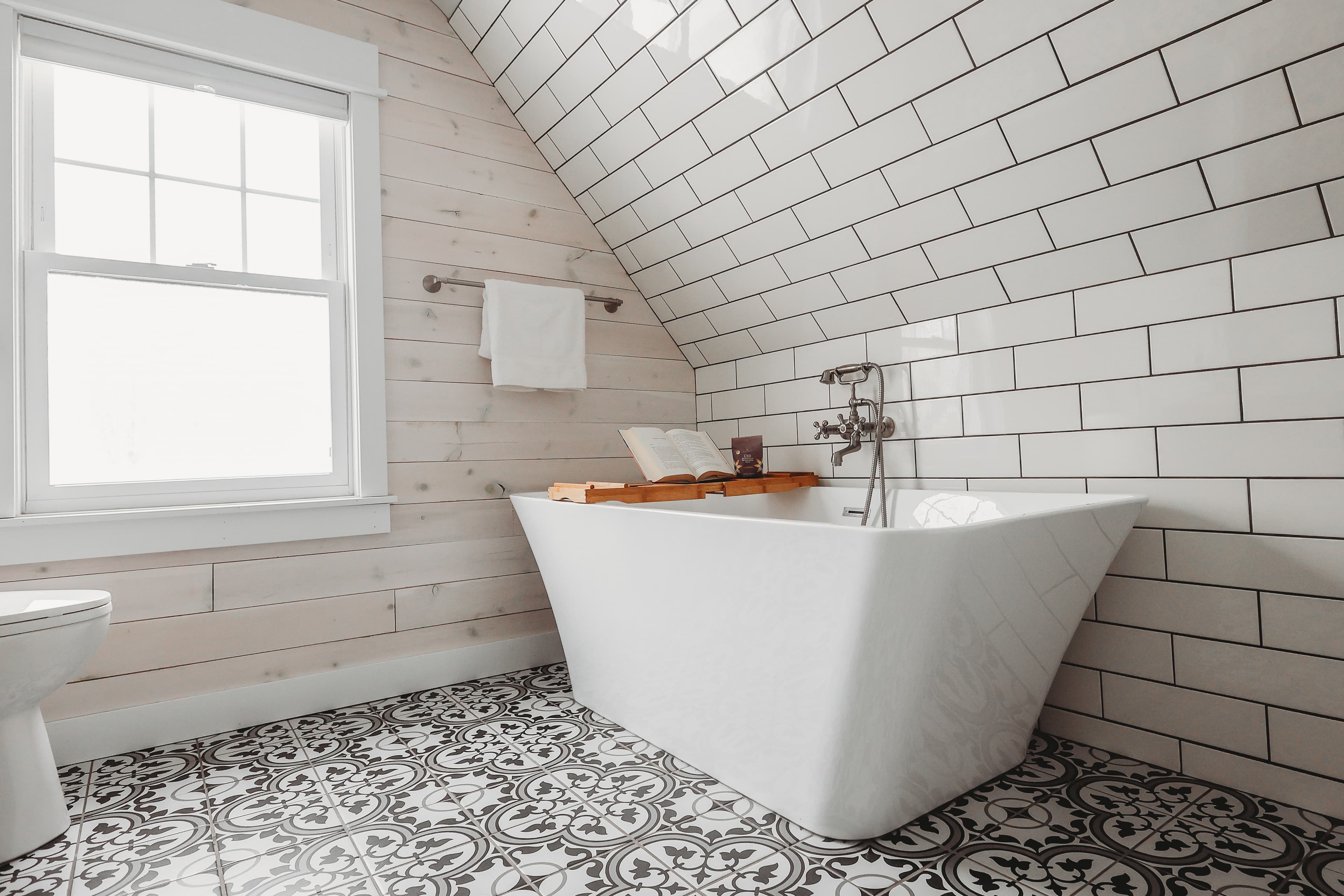 Modern bathroom with a white freestanding tub, sloped subway tile wall, and patterned floor tiles.