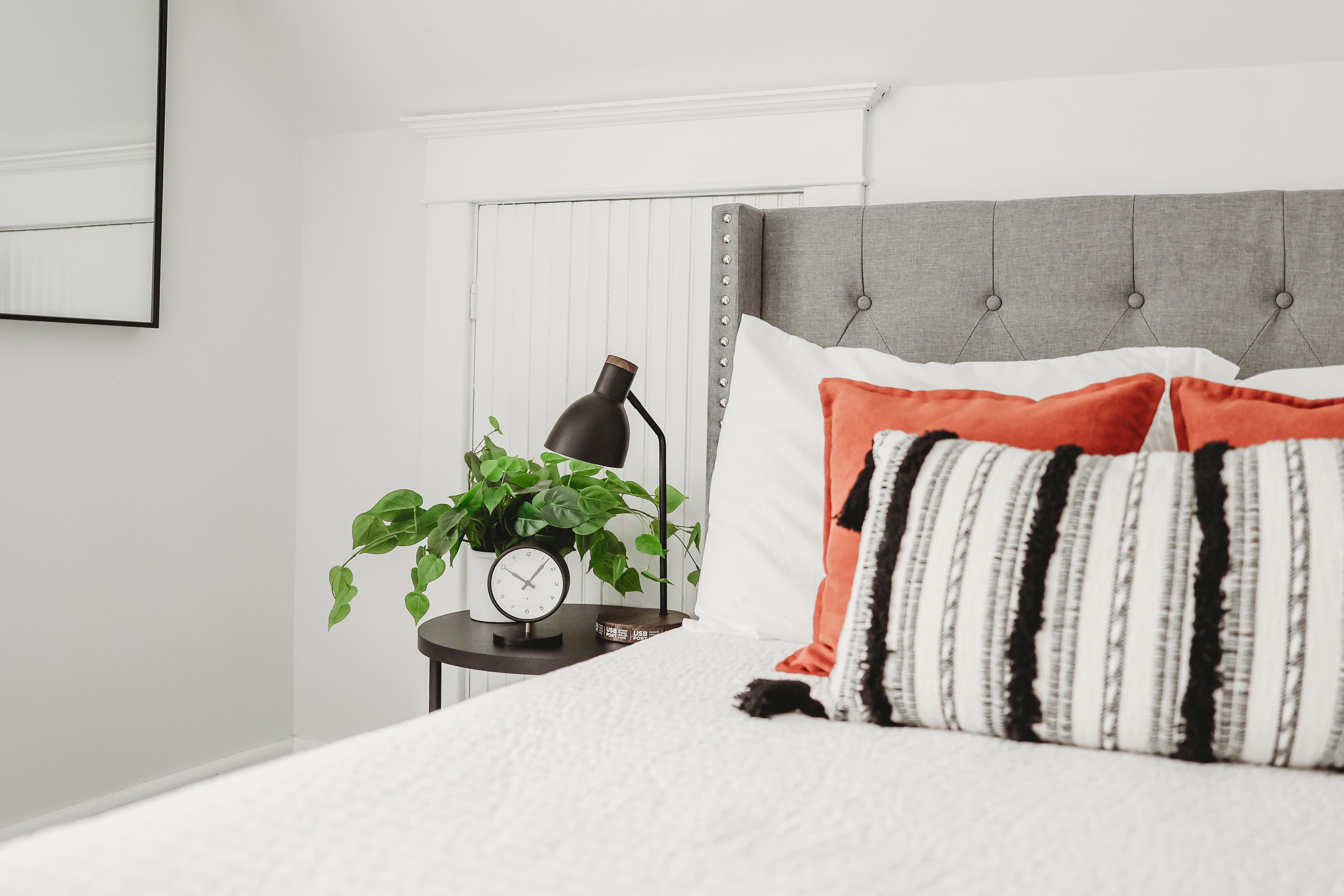 Modern bedroom with a grey tufted headboard, orange pillows, and a plant on the nightstand.