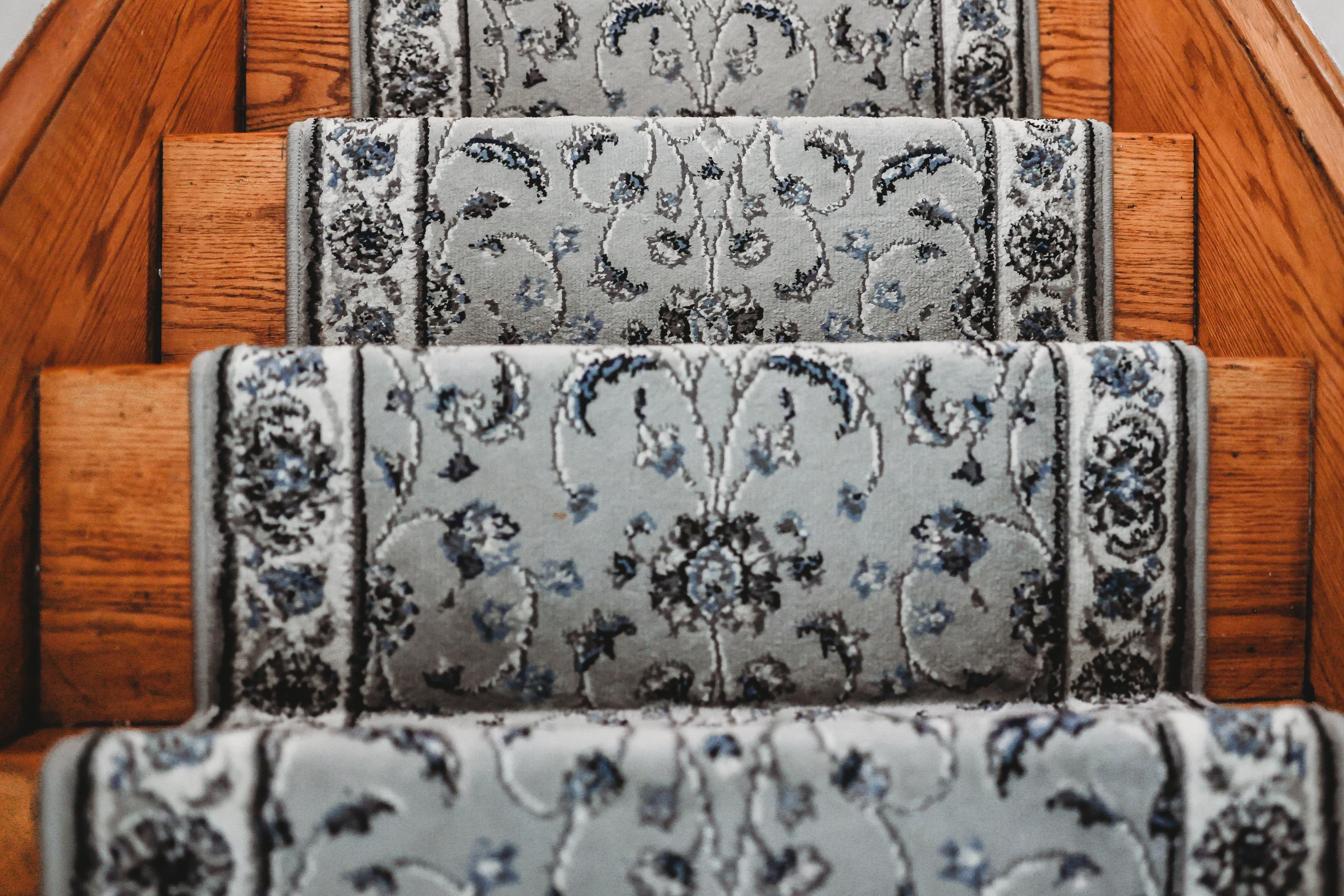 Wooden staircase steps covered with a grey and blue ornate floral patterned carpet runner.