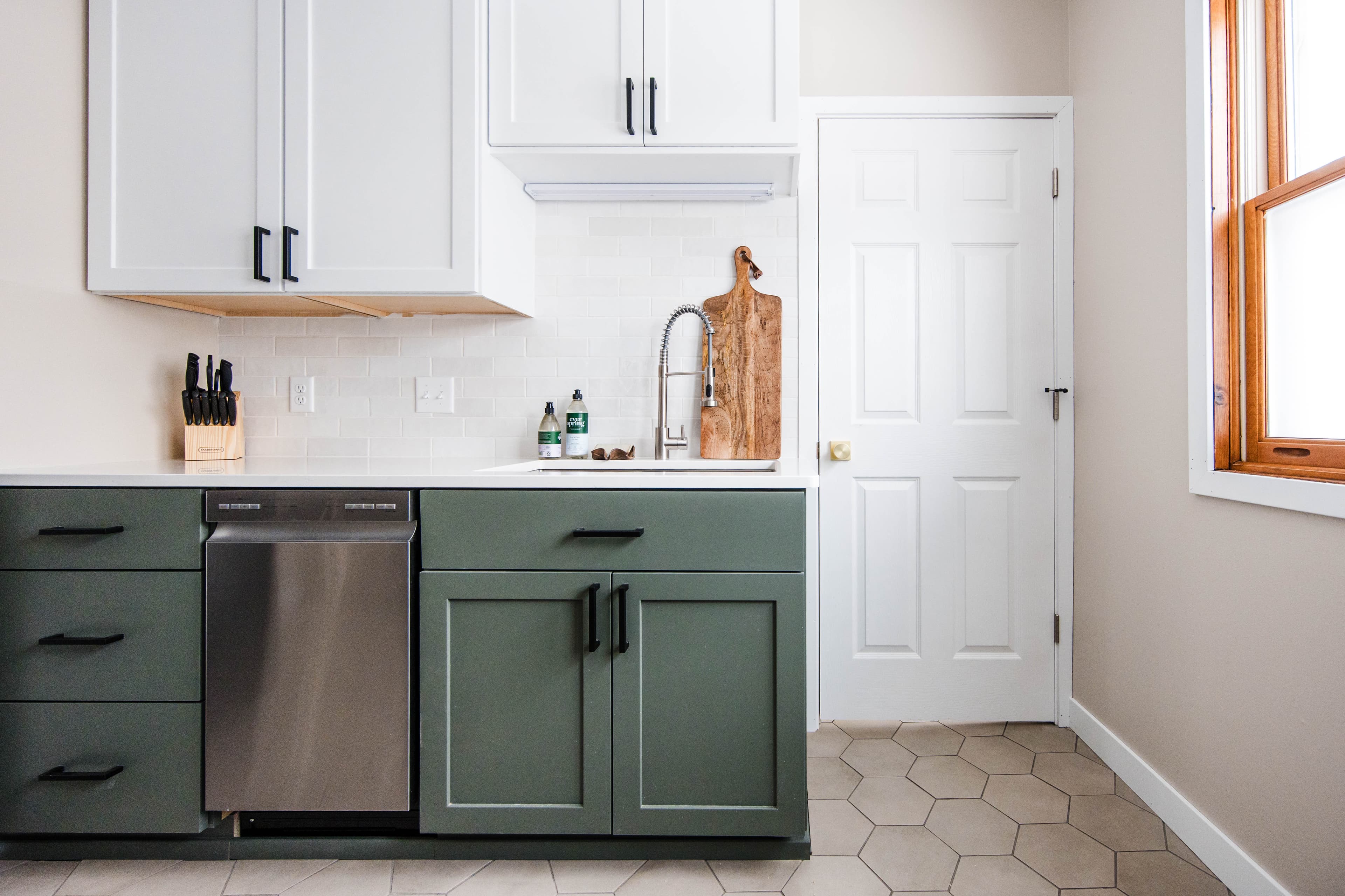 Modern kitchen with sage green and white cabinets, stainless dishwasher, and hexagonal floor tiles.