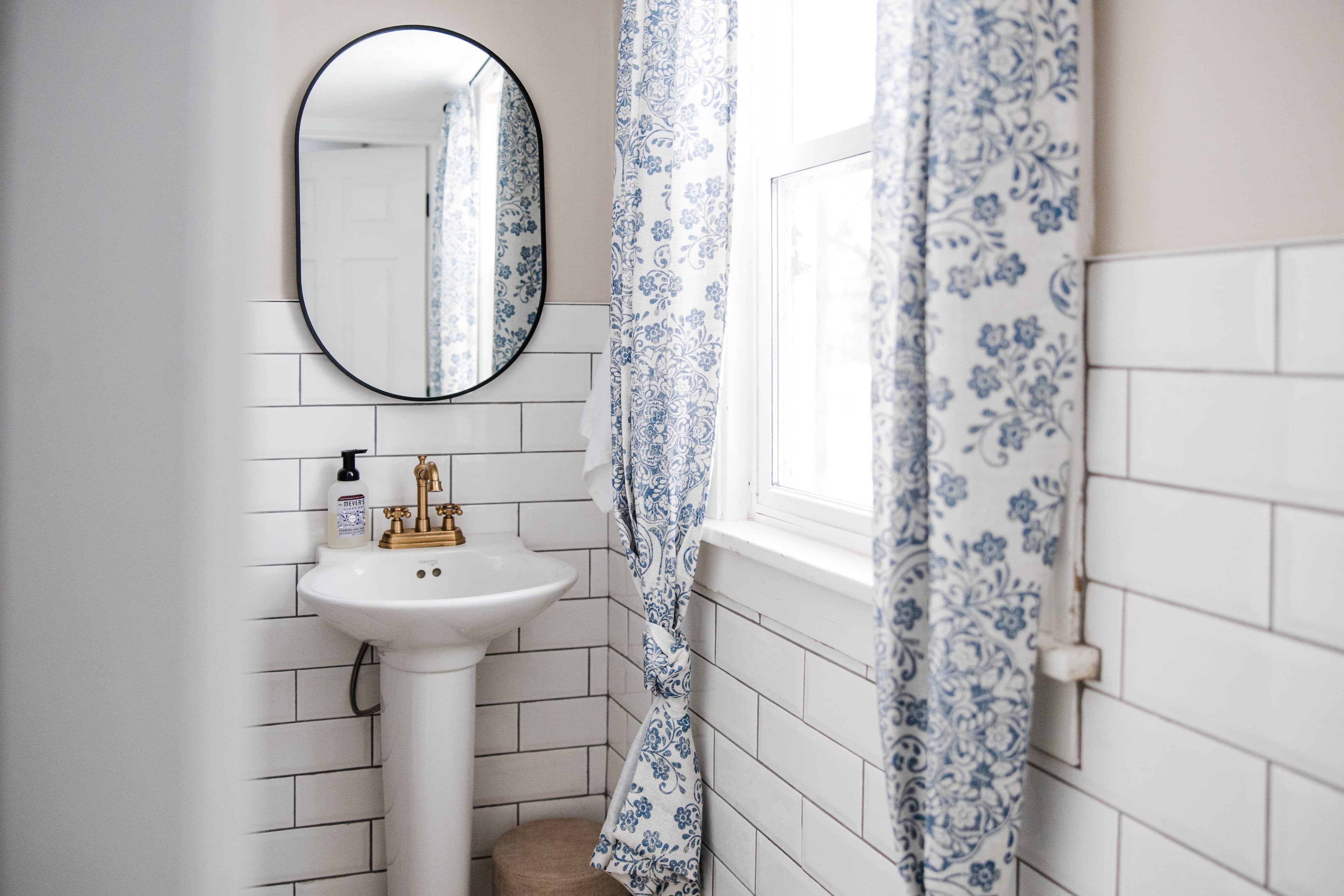 White pedestal sink with gold faucet, oval mirror, and blue floral curtains against subway tile.