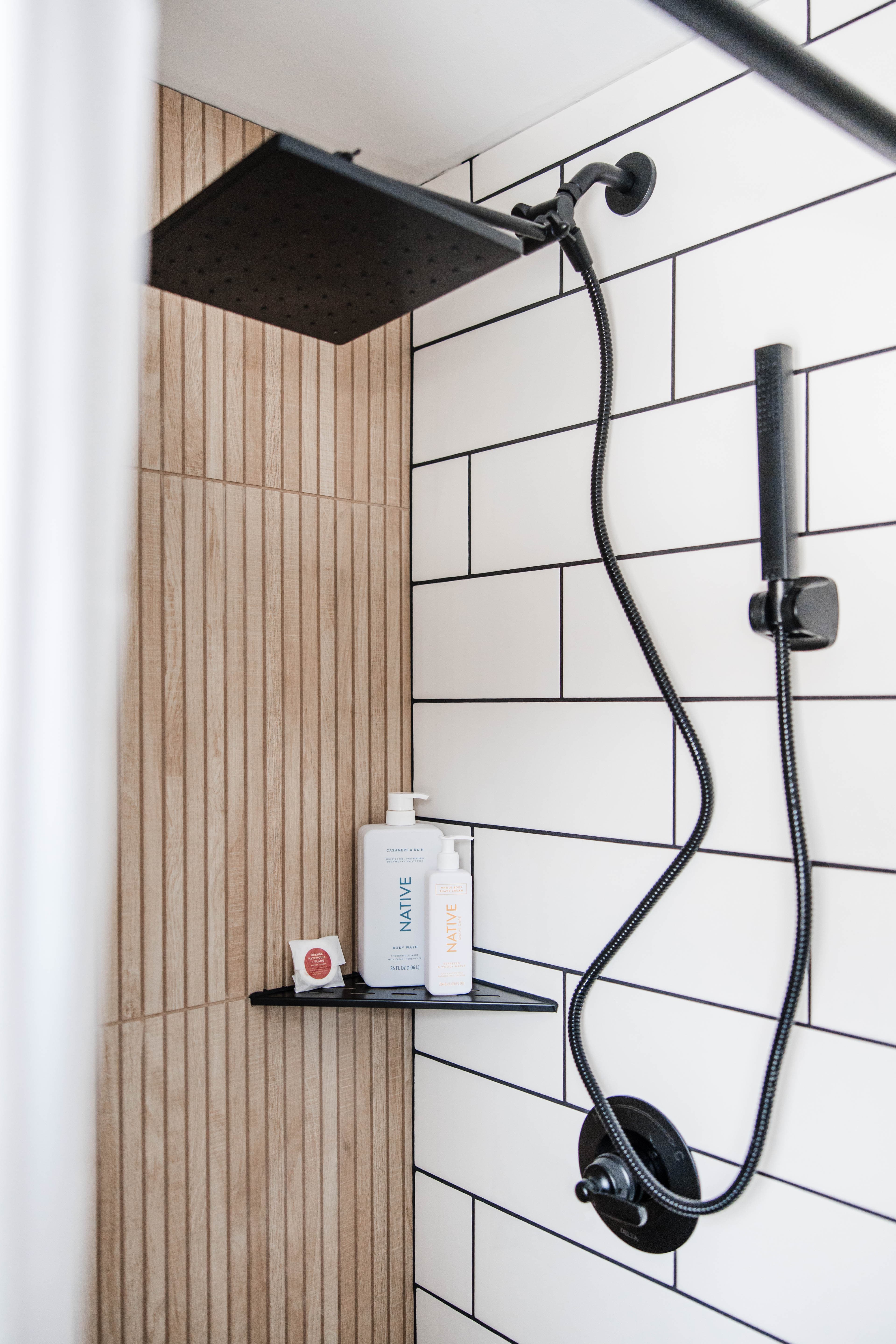 Modern shower with matte black fixtures, white subway tile, wood-look accents, and Native toiletries.