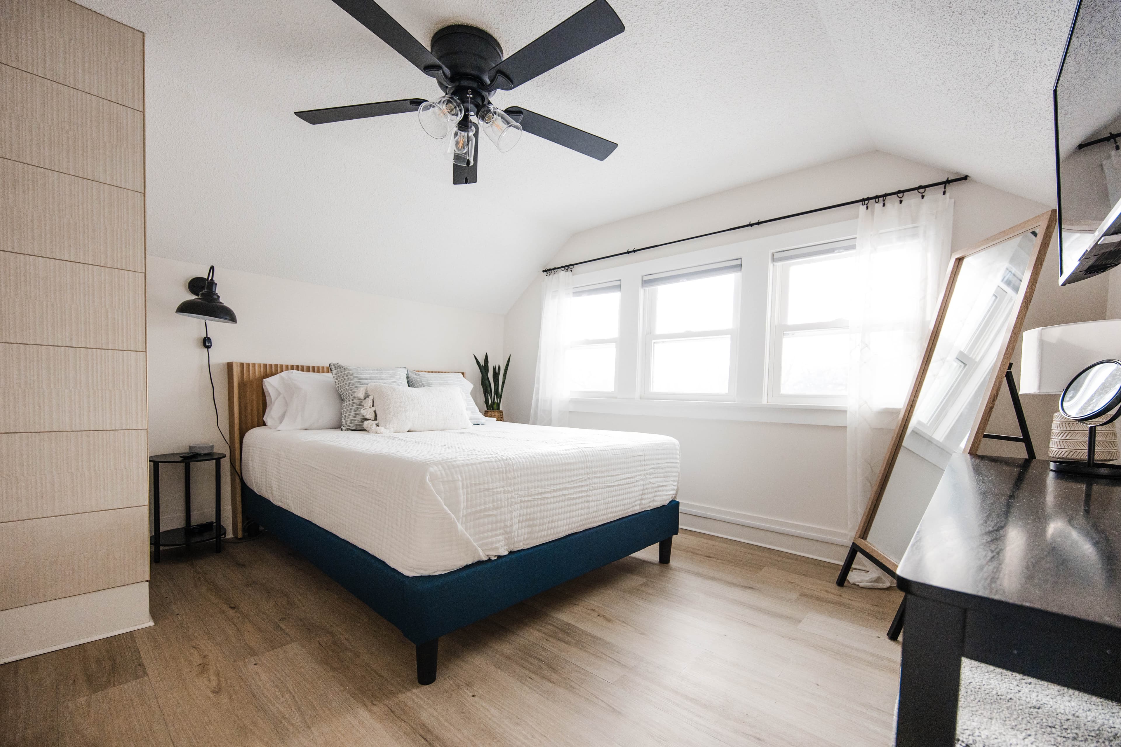 Bright bedroom featuring a blue bed, white bedding, large windows, and a black ceiling fan.