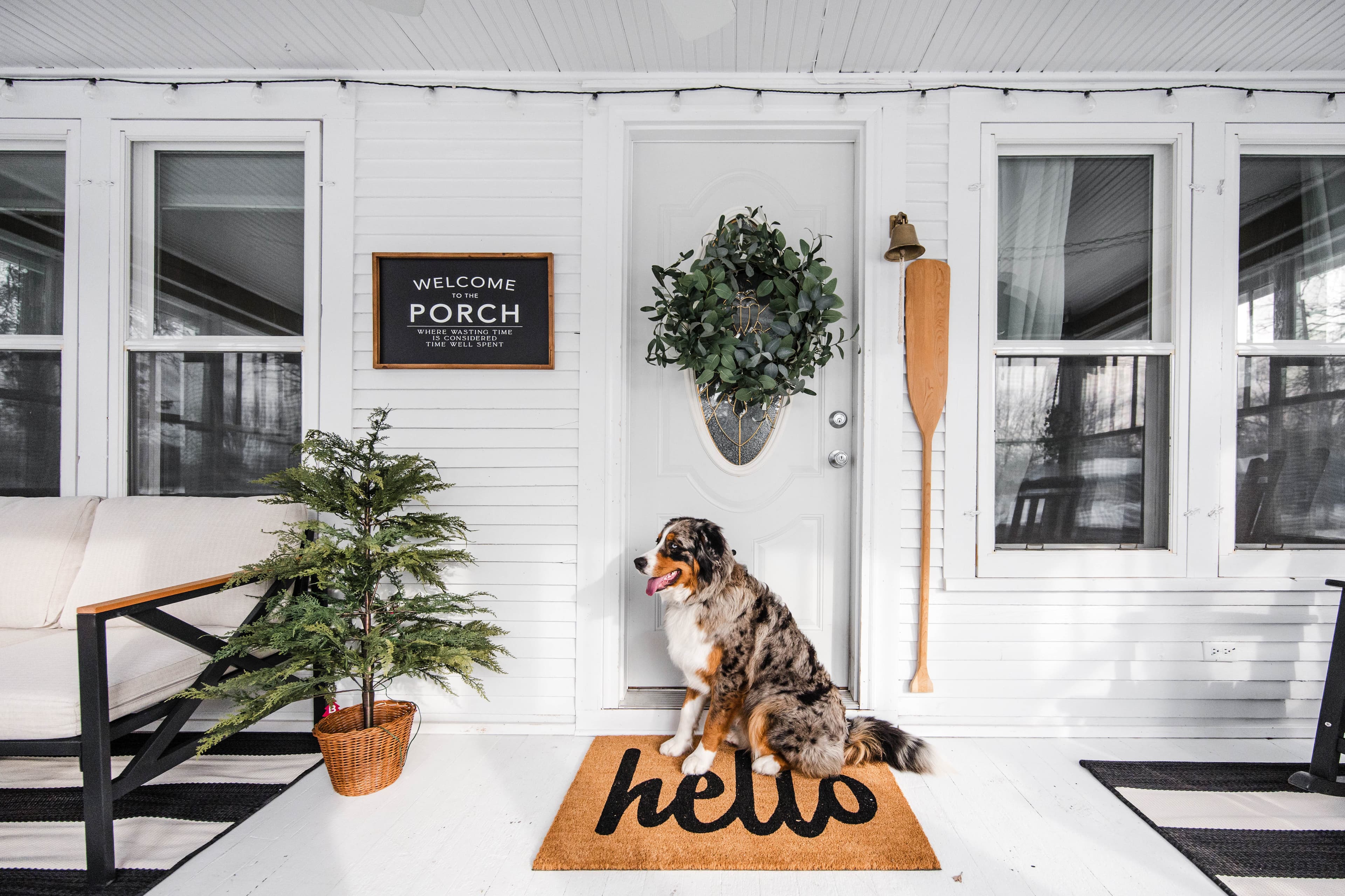 Merle Australian Shepherd sitting on a hello doormat on a decorated white front porch.
