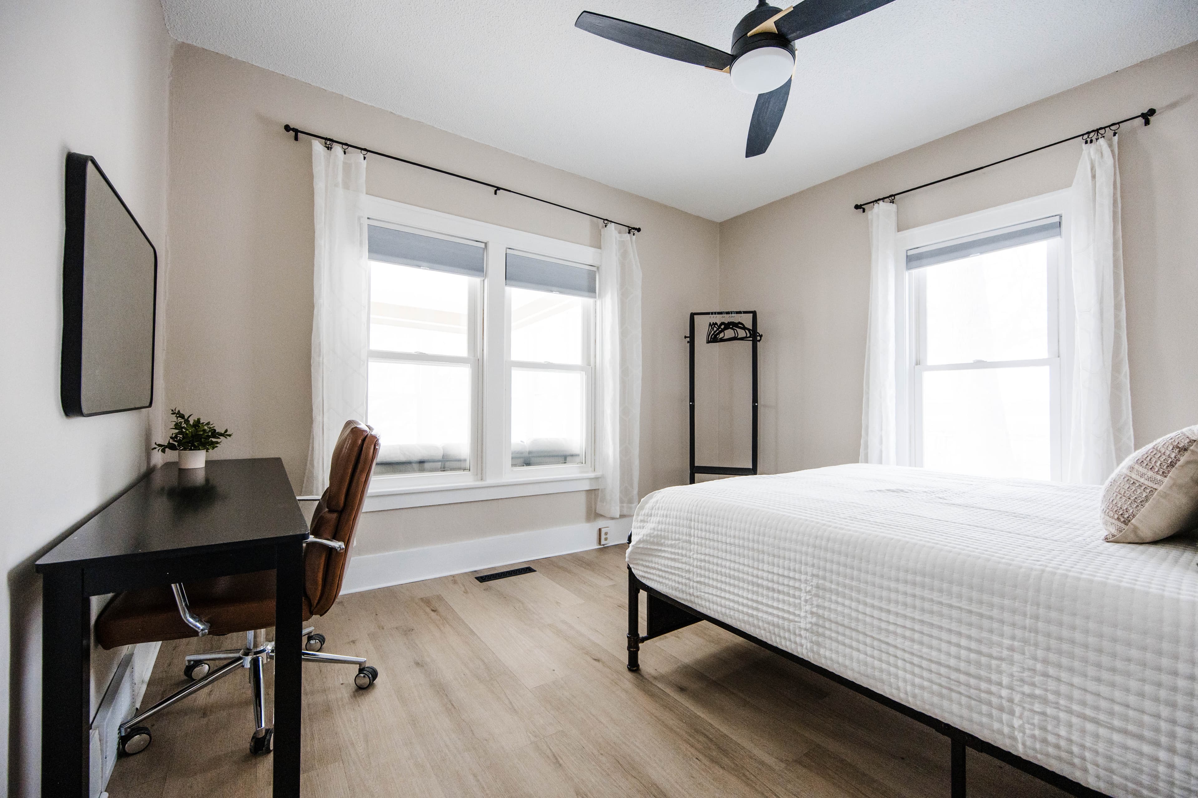 Modern bedroom with a white bed, black desk, brown leather chair, and light wood floors.