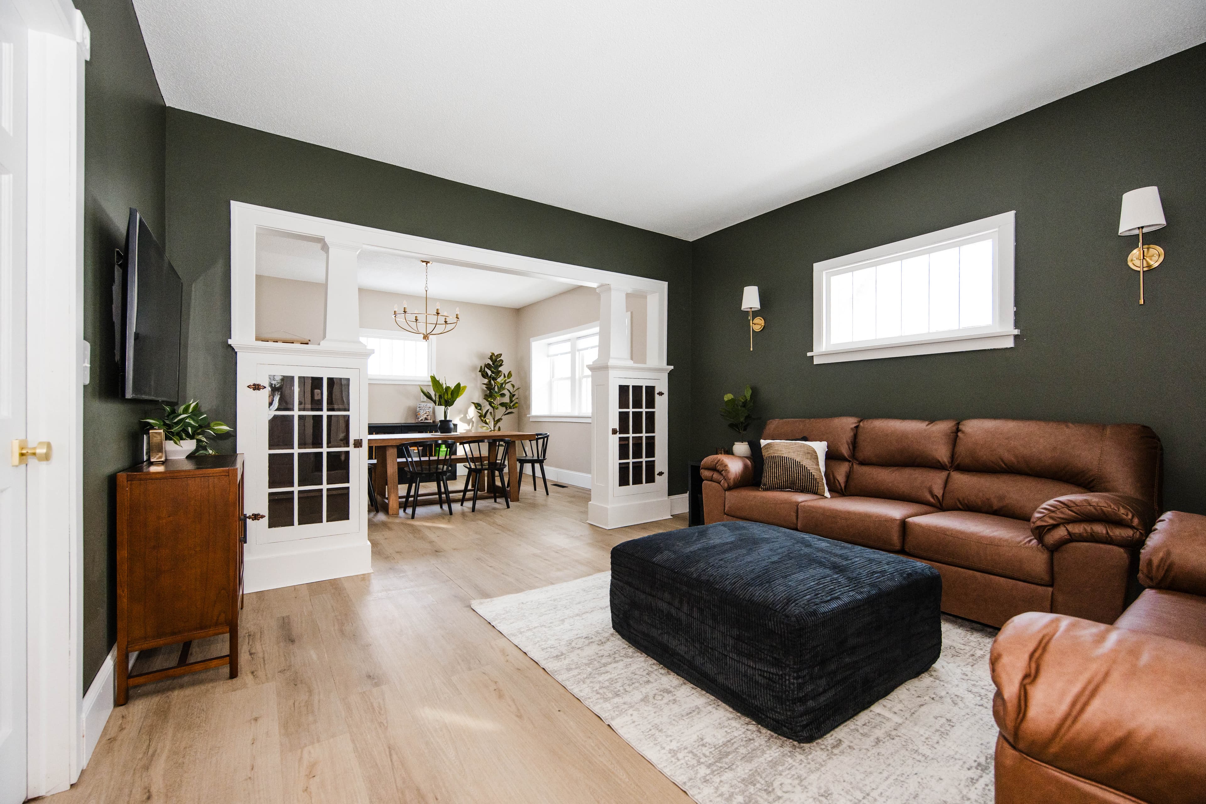 Dark green living room with brown leather sofa, blue ottoman, and view into dining room.